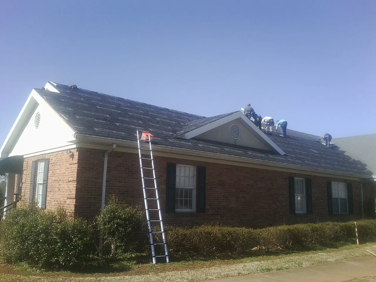 Crew tearing shingles off a brick ranch roof with a ladder set — Emergency Roof Repair work by our crew in Iola